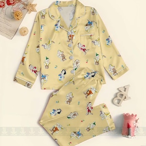 May include: Yellow pajama set featuring a print of Snow White and the Seven Dwarfs. The long-sleeved top has a button-down front and white trim. The matching pants have an elastic waistband. The set is made of a soft, silky material.
