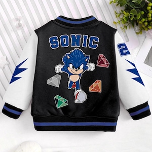 May include: Black and white varsity jacket featuring the character Sonic the Hedgehog. The jacket has blue and white sleeves with lightning bolt designs. The word "SONIC" is printed across the back in blue letters, along with a graphic of Sonic and colorful gems.