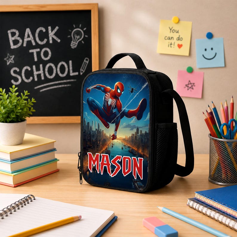 Puede incluir: Una bolsa de almuerzo negra con un gr&aacute;fico de un superh&eacute;roe rojo y azul saltando sobre el horizonte de una ciudad. El nombre MASON est&aacute; impreso en letras rojas grandes y negritas en la parte inferior. La bolsa tiene un asa superior y una cremallera.