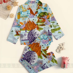 May include: Light blue pajama set featuring a whimsical print of a purple dragon, mountains, and architectural elements. The long-sleeved top has white trim and buttons. The matching shorts have the same print. A fun and colorful sleepwear set.