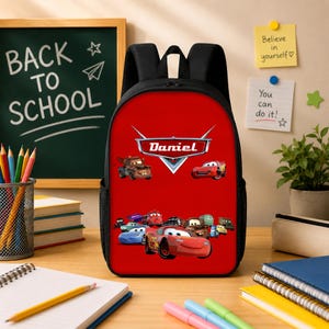 May include: A black backpack with a bright red front panel featuring a graphic of various animated race cars and the name Daniel printed in a stylised silver emblem. The backpack has black shoulder straps and side mesh pockets for storage.