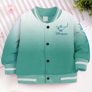May include: A green and white ombre bomber jacket with a white ribbed collar and cuffs. The jacket features a blue and white graphic of a genie lamp and the text "Disneyland Paris".
