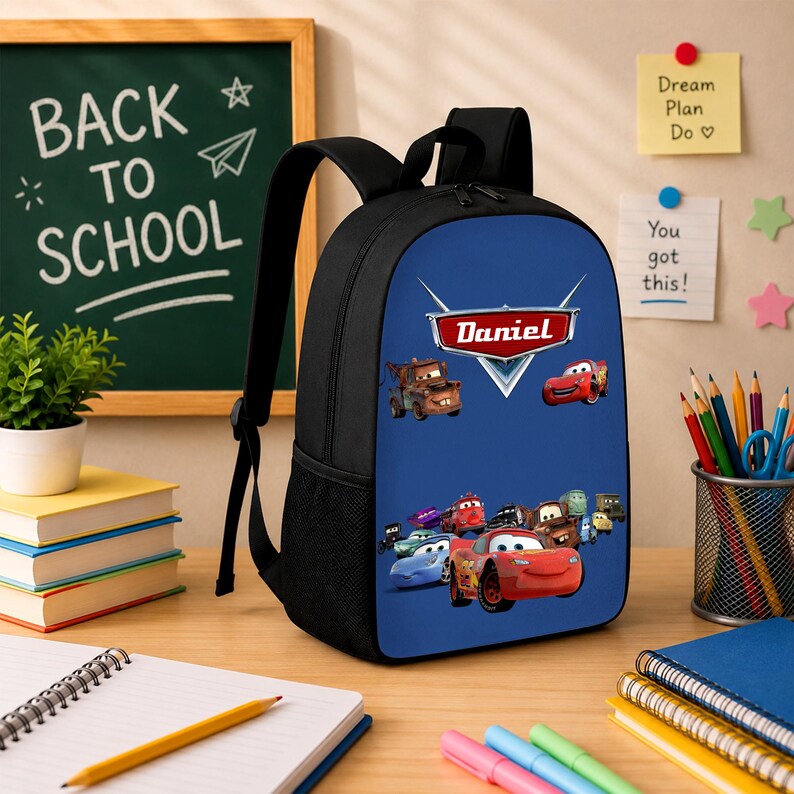 May include: A black backpack with a blue front panel featuring a graphic print of various animated car characters and a red emblem with the name Daniel printed in white text. The backpack has adjustable shoulder straps and side mesh pockets.