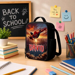 May include: A black lunch bag featuring a graphic of Spider-Man leaping over a city skyline at sunset. The name DAVID is printed in large, bold, red and white letters below the character. The bag has a top handle and a side mesh pocket.