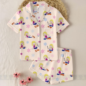 Pu&ograve; includere: Un completo pigiama a due pezzi beige chiaro, con un motivo ripetuto di personaggi dei cartoni animati e fiori. Il set comprende una camicia a maniche corte con bottoni e pantaloncini abbinati, entrambi bordati da un profilo bianco.