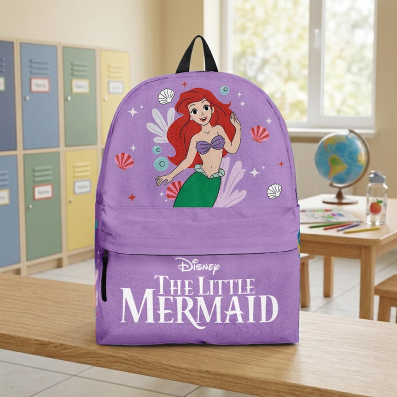 May include: A purple backpack featuring Ariel from Disney's The Little Mermaid. The backpack has a large image of Ariel, with red hair and a green tail, on the front. The words "The Little Mermaid" are printed in white on the lower section.