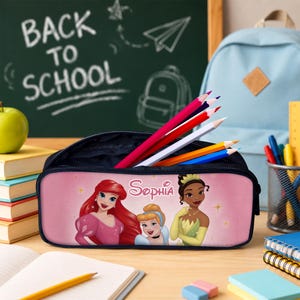 May include: A rectangular pink pencil case featuring illustrations of three animated princesses and the name Sophia printed in white script. The case is open and contains several coloured pencils, with a light blue backpack and books nearby.