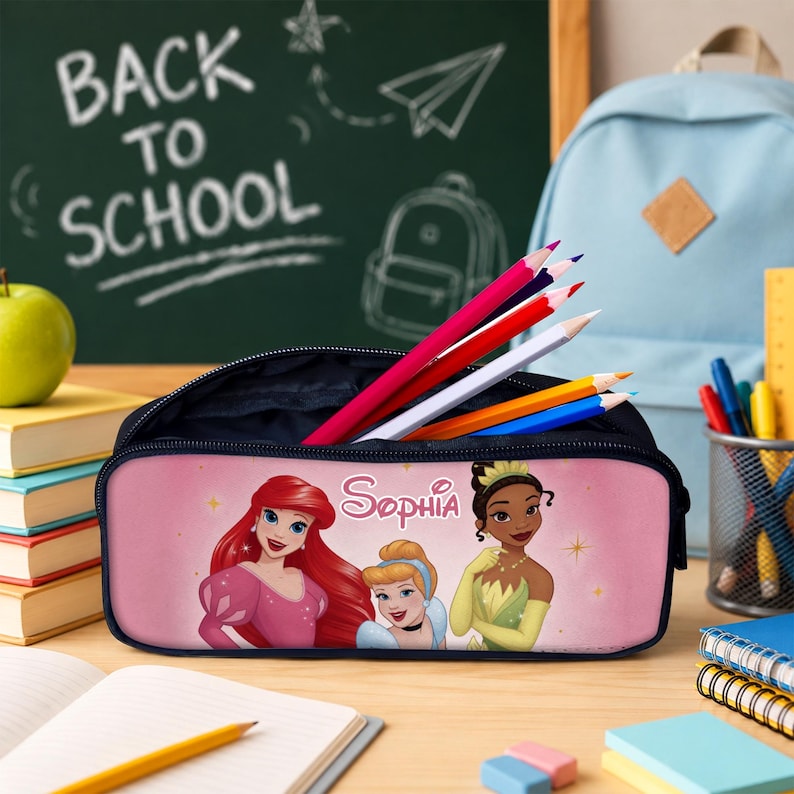May include: A rectangular pink pencil case featuring illustrations of three animated princesses and the name Sophia printed in white script. The case is open and contains several coloured pencils, with a light blue backpack and books nearby.