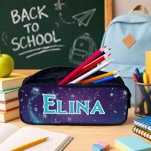 May include: A dark blue pencil case featuring a starry night sky pattern with white snowflakes and the name ELINA printed in large, light blue letters. The case is open and contains several colored pencils in red, white, yellow, and blue.