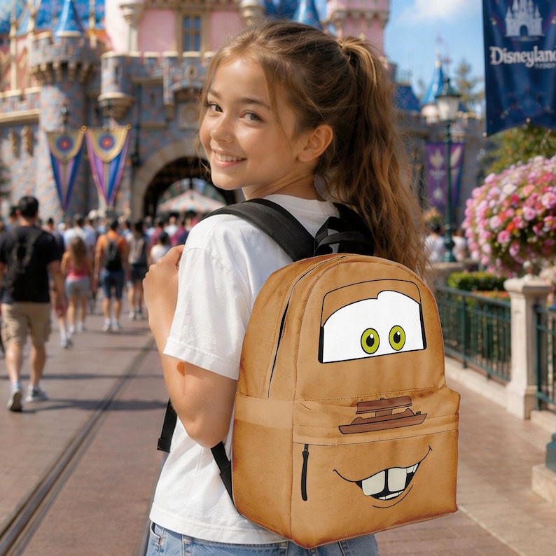Puede incluir: Una mochila marr&oacute;n que presenta al personaje animado Mate de la pel&iacute;cula Cars. La mochila tiene un bolsillo frontal grande y un cierre de cremallera. El dise&ntilde;o incluye los ojos, la boca y la parte delantera de Mate. El fondo muestra un parque tem&aacute;tico.