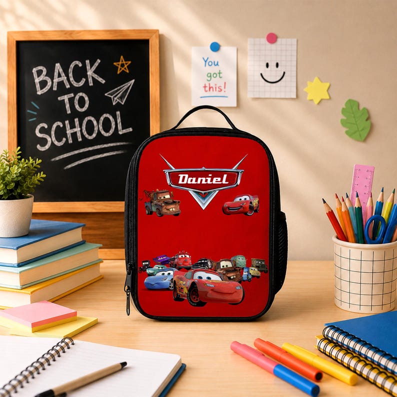 May include: A red rectangular lunch bag featuring the Disney Pixar Cars characters and the name Daniel printed in white text on a silver emblem. The bag has a black handle, black trim, and a side mesh pocket, set against a bright red background.