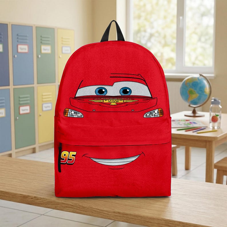 Puede incluir: Una mochila roja con la cara de un coche de dibujos animados, con ojos azules y una boca sonriente. El n&uacute;mero "95" est&aacute; impreso en la parte inferior izquierda. Tiene un asa negra y es adecuada para la escuela o los viajes.