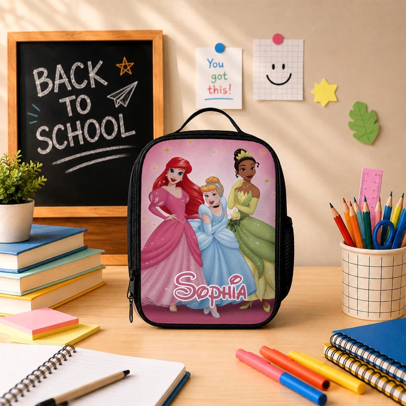 May include: A black lunch bag featuring a pink background with illustrations of Ariel, Cinderella, and Tiana. The name Sophia is printed in white script at the bottom centre. The bag has a black zip, a top handle, and a side mesh pocket.