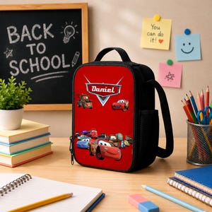 May include: A rectangular black lunch bag with a red front panel featuring a graphic print of various Cars characters and the name Daniel in a white logo emblem. The bag has a top handle, a side mesh pocket, and a zipper closure along the perimeter.