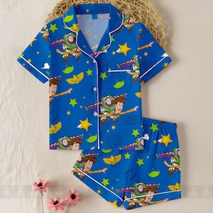 May include: Blue pajama set featuring a print of Woody and Buzz Lightyear from Toy Story, with stars and other graphics. The short-sleeved top and shorts have white trim.