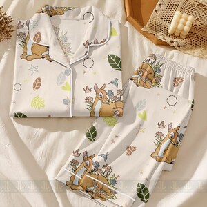 Op de afbeelding: Cr&egrave;mekleurige pyjamaset met een Bambi en Stampertje print. De top met lange mouwen heeft een knoopsluiting en witte bies. De broek heeft een elastische tailleband. De set bevat illustraties van bloemen, bladeren en vogels.