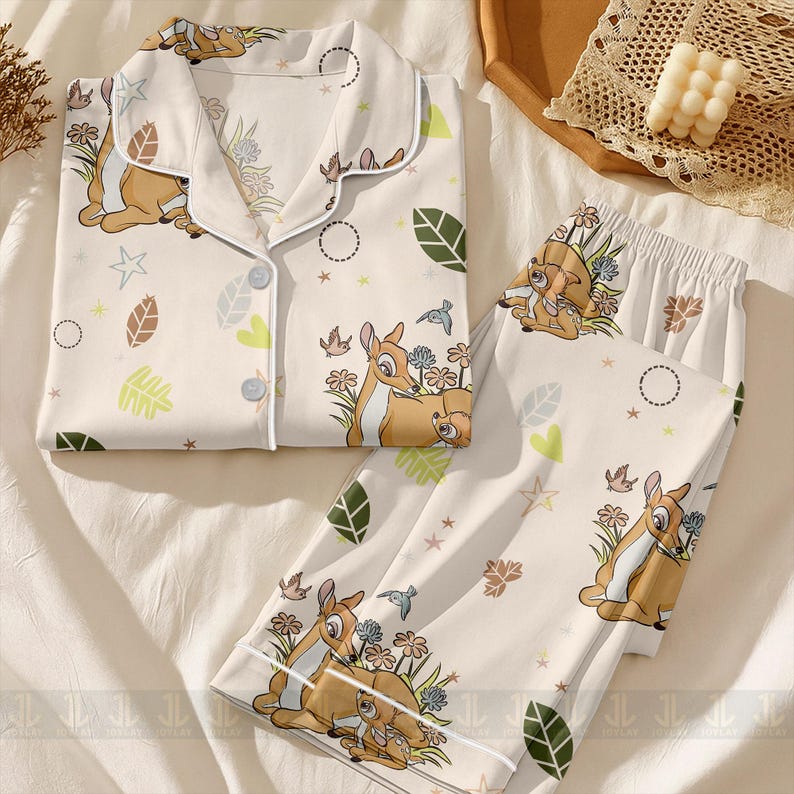 Op de afbeelding: Cr&egrave;mekleurige pyjamaset met een Bambi en Stampertje print. De top met lange mouwen heeft een knoopsluiting en witte bies. De broek heeft een elastische tailleband. De set bevat illustraties van bloemen, bladeren en vogels.