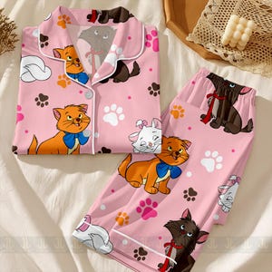 May include: Pink pajama set featuring cartoon cats, paw prints, and clouds. The top has a button-down front with white trim. The pants and top are covered in illustrations of orange, white, and brown cats with blue bows and red ribbons.