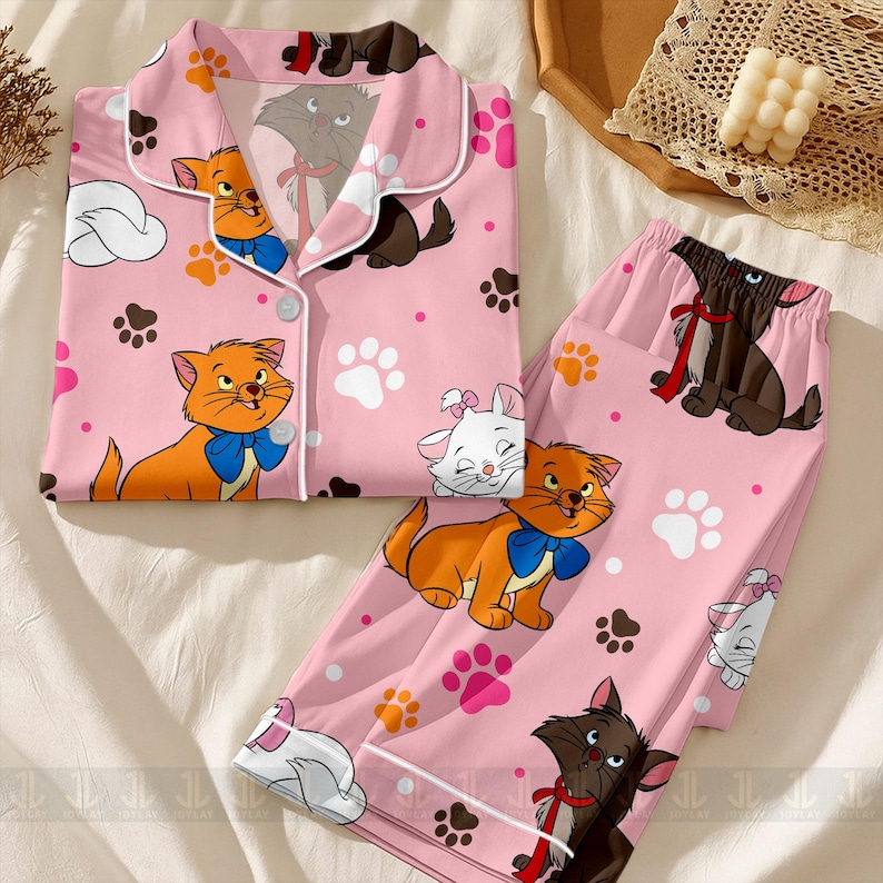 May include: Pink pajama set featuring cartoon cats, paw prints, and clouds. The top has a button-down front with white trim. The pants and top are covered in illustrations of orange, white, and brown cats with blue bows and red ribbons.