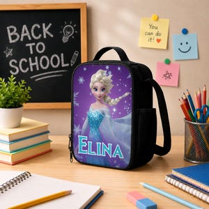 May include: A black lunch bag featuring a graphic of a blonde character in a blue dress with the name ELINA printed in light blue letters. The bag has a top handle, a zipper closure, and a side mesh pocket for storage on a desk surface.