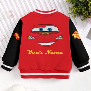 May include: Red and black baseball jacket with a cartoon image of a red race car with a smiling face. The jacket has white stripes on the sleeves and bottom. The text "Your Name" is printed in yellow on the front of the jacket.