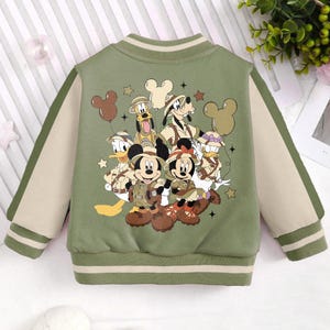 May include: A green and beige bomber jacket with a colourful cartoon print of Mickey Mouse and his friends. The print features Mickey Mouse, Minnie Mouse, Donald Duck, Goofy, and Pluto in safari outfits.
