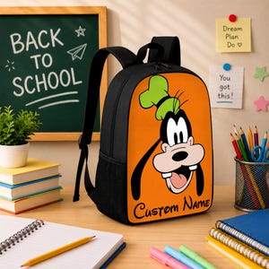 May include: A black backpack featuring a large orange front panel with a graphic of the cartoon character Goofy wearing a green hat. The text "Custom Name" is printed in black script at the bottom of the orange panel. The bag has black straps.