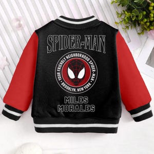 May include: Black and red baseball jacket with a Spider-Man logo on the back. The logo features a red and black Spider-Man mask with the text "Your Friendly Neighborhood Spider-Man - Brooklyn, New York". The text "Miles Morales" is printed below the logo.