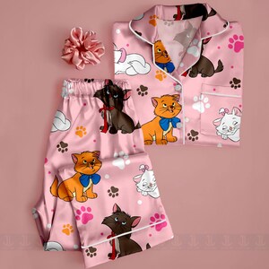 May include: Pink pajama set with a matching scrunchie. The pajamas feature a print of cartoon cats, paw prints, and clouds. The set includes a button-down top and pants with white trim.