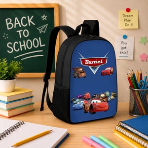 K&ouml;nnte beinhalten: Ein schwarzer Rucksack mit einer blauen Frontpartie, die einen Grafikdruck verschiedener animierter Autofiguren sowie ein rotes Emblem mit dem in Wei&szlig; gedruckten Namen Daniel zeigt. Der Rucksack verf&uuml;gt &uuml;ber verstellbare Schultergurte und seitliche Netztaschen.