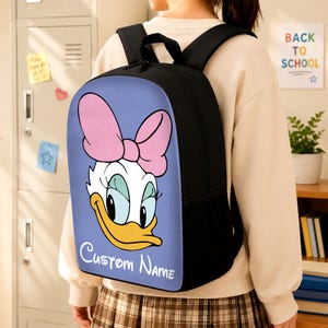 May include: A black backpack featuring a light purple front panel with a graphic of Daisy Duck wearing a large pink bow. The character has white feathers, long eyelashes, and a yellow beak. The text Custom Name is printed in white at the bottom.