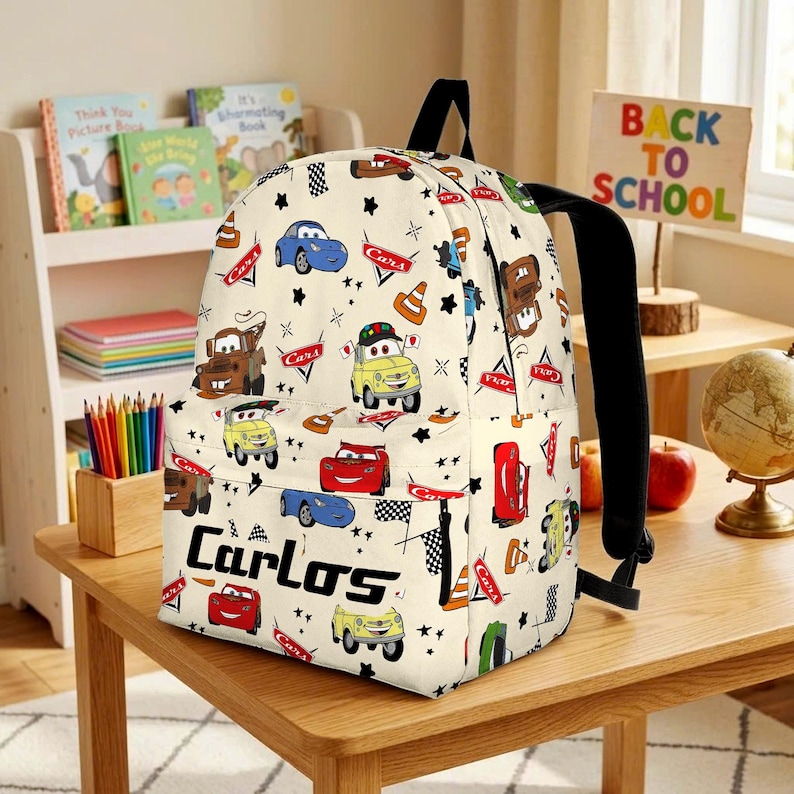 May include: A cream-coloured backpack featuring a repeating pattern of various cartoon cars, traffic cones, and checkered flags. The name Carlos is printed in bold black letters on the front pocket, accompanied by small black star shapes.