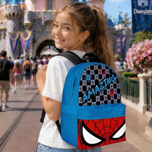 May include: A blue backpack featuring a Spider-Man design. The backpack has a red and black spiderweb design on the bottom and a checkered pattern with the word "AMAZING" on the top. The backpack is being worn by a person.