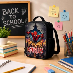 May include: A black rectangular lunch bag featuring a graphic of a red superhero emerging from a brick wall above a city skyline. The name CARLOS is printed in bold red and white letters below the superhero. The bag has a top handle and side mesh.