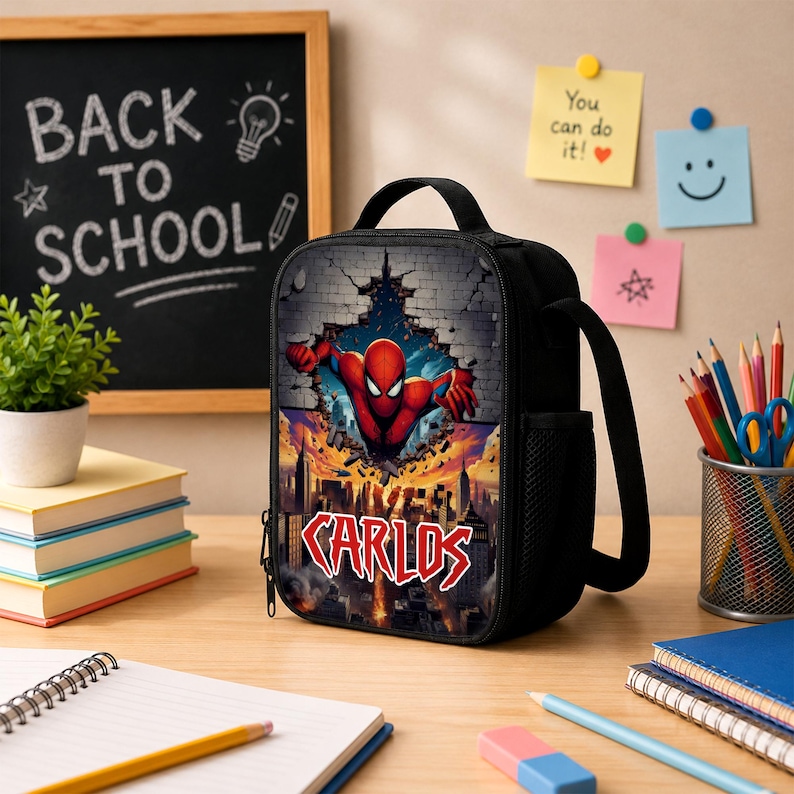 May include: A black rectangular lunch bag featuring a graphic of a red superhero emerging from a brick wall above a city skyline. The name CARLOS is printed in bold red and white letters below the superhero. The bag has a top handle and side mesh.