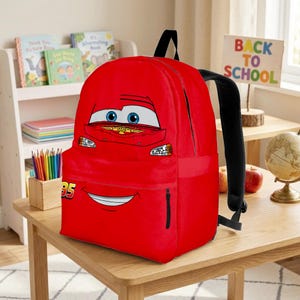 Puede incluir: Una mochila roja brillante con el personaje de Rayo McQueen de la pel&iacute;cula Cars. La mochila tiene una cremallera negra, correas negras y el n&uacute;mero 95. Un letrero de "Back to School" es visible en el fondo.