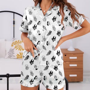 K&ouml;nnte beinhalten: Wei&szlig;es Satin-Pyjama-Set mit einem sich wiederholenden Muster von Cartoon-B&ouml;sewicht-Gesichtern in Schwarz und Grau. Das Set beinhaltet ein kurz&auml;rmeliges Hemd mit Kn&ouml;pfen und passende Shorts. Der Pyjama hat wei&szlig;e Paspeln.