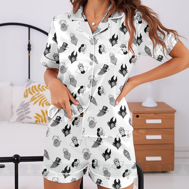 K&ouml;nnte beinhalten: Wei&szlig;es Satin-Pyjama-Set mit einem sich wiederholenden Muster von Cartoon-B&ouml;sewicht-Gesichtern in Schwarz und Grau. Das Set beinhaltet ein kurz&auml;rmeliges Hemd mit Kn&ouml;pfen und passende Shorts. Der Pyjama hat wei&szlig;e Paspeln.