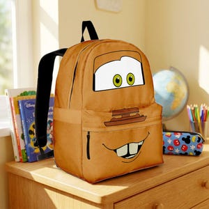 May include: A tan backpack featuring a graphic of a cartoon character face with large white eyes, green pupils, and a wide, toothy grin. The bag has black shoulder straps, a main zippered compartment, and a smaller front pocket with a black zipper.