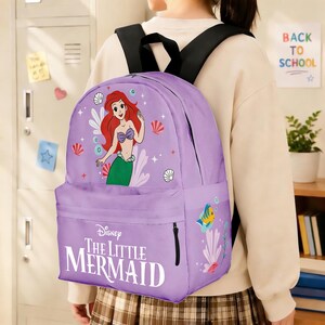May include: A purple backpack featuring Ariel from Disney's The Little Mermaid. The backpack has a front pocket and black shoulder straps. The design includes Ariel, seashells, and the movie title.