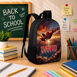 May include: A black backpack featuring a graphic print of a red and blue superhero flying over a city skyline at sunset. The name DAVID is printed in large red letters across the center of the image, positioned above a pile of broken bricks.