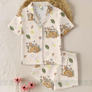 Op de afbeelding: Cr&egrave;mekleurige pyjamaset met een Bambi en Stampertje print. De top met korte mouwen heeft een reverskraag en een voorzak. De shorts hebben een elastische tailleband. De set is versierd met bladeren, bloemen en sterren.