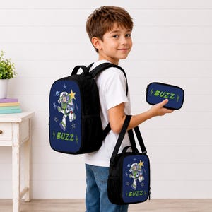May include: A black backpack, a matching lunch bag, and a small pencil case, all featuring a blue front panel with a graphic of Buzz Lightyear, yellow stars, and the text BUZZ in green. The items have black straps and side mesh pockets.