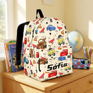 May include: A cream-coloured backpack featuring a repeating pattern of characters and icons from the movie Cars, including race cars, tow trucks, traffic cones, and checkered flags. The name Sofia is printed in black text on the front pocket.