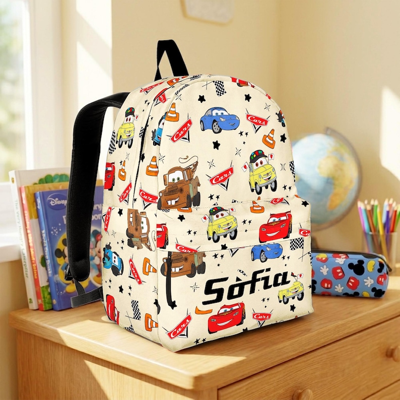 May include: A cream-coloured backpack featuring a repeating pattern of characters and icons from the movie Cars, including race cars, tow trucks, traffic cones, and checkered flags. The name Sofia is printed in black text on the front pocket.