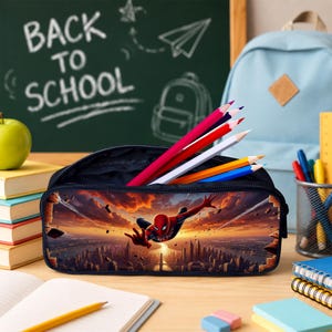 May include: A black zippered pencil case featuring a graphic print of a superhero in a red and blue suit flying over a city skyline at sunset. The case is filled with several colored pencils, including red, white, yellow, and blue shades.