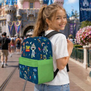 May include: A navy blue backpack featuring characters from Toy Story, with a green front pocket. The backpack has a black back and straps. The words "Toy Story" are printed on the front. The background shows a theme park.