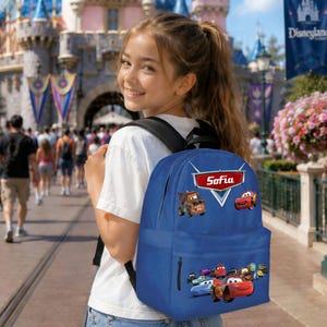 May include: A blue backpack with characters from the film Cars, and the name "Sofia" on a red emblem. The backpack has a front zip pocket. The person wearing a white t-shirt is in a theme park.