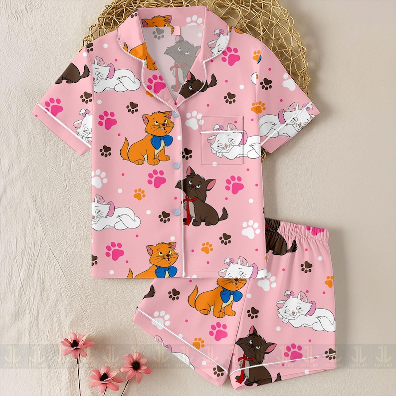 May include: Pink pajama set featuring a pattern of cartoon cats, paw prints, and polka dots. The short-sleeved top has a button-down front and a chest pocket. The shorts have an elastic waistband and white trim.