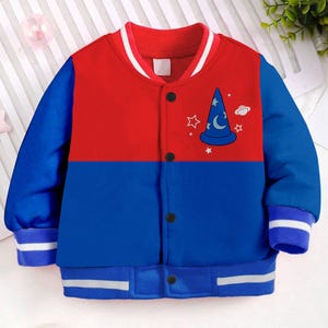 May include: A red and blue baseball jacket with a white stripe on the collar and cuffs. The jacket has a cartoon image of a wizard's hat with stars and a crescent moon on the front.